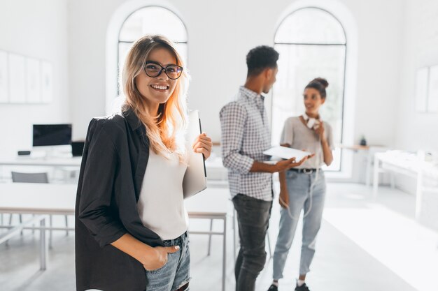 Elegant business-lady in trendy black jacket holding laptop and smiling. Portrait of cheerful blonde secretary and tall african office worker.