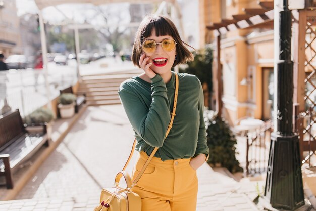都会の春の日に幸せを表現するサングラスでエレガントな白人の女の子。笑っている黄色のハンドバッグと素敵な女性モデルの屋外写真