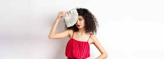 Free photo elegant curlyhaired woman in red dress showing fan of money and looking confident rich girl holding