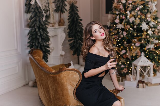 Elegant, expensive New Year's atmosphere in apartment contributes to wonderful mood of elegant, attractive brunette with bright lips, holding glass of sparkling wine