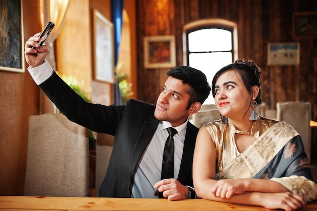Elegant and fashionable indian friends couple of woman in saree and man in suit posed indoor cafe and making selfie on mobile phone