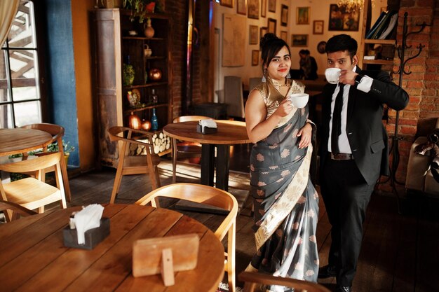 Elegant and fashionable indian friends couple of woman in saree and man in suit sitting on cafe and drinking tea