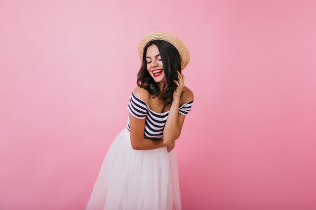 Elegant female model with red lips posing in white skirt. Indoor shot of amazing brunette woman in straw hat standing.