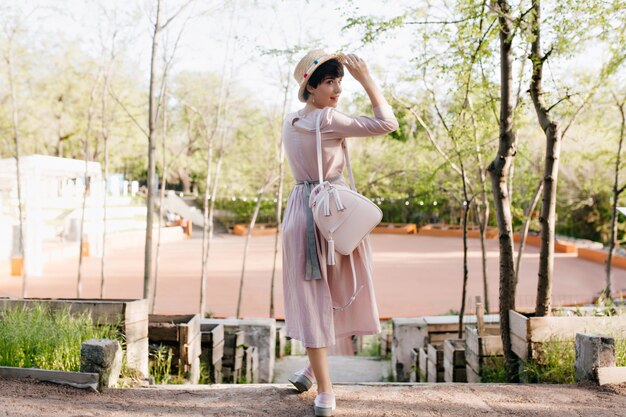 Elegant girl in old-fashioned attire looking over her shoulder and holding straw hat with cute smile