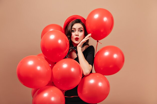 Elegant girl in red beret and black dress blows kiss and holds huge balloons.