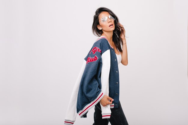 Elegant girl wears big trendy glasses posing and playing with shiny hair. European young woman in stylish accessories holding blue sport jacket isolated.