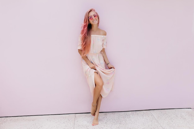 Элегантная девушка с загорелой кожей играет со своим длинным платьем на белом фоне Смеющаяся потрясающая женщина с розовыми волосами веселится во время фотосессии в помещении