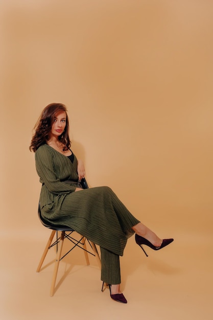 Elegant incredible woman with dark hair is dressed dark green suit and heels and sitting on chair over isolated background