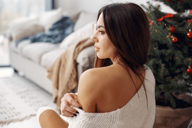 Free photo elegant lady near christmass tree. woman in a room. famale in a white knited sweater.