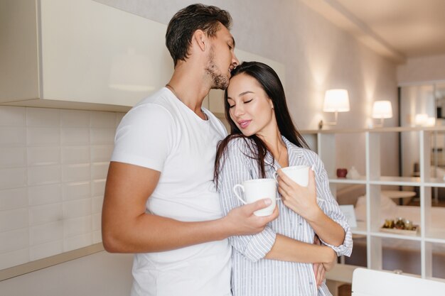 Elegant lady wears striped pajama hugs her boyfriend holding cup of coffee