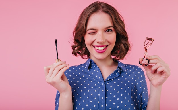 Elegant lady with short hairstyle doing her eye lashes and laughing. Indoor photo of smiling curly woman holding mascara on pink wall.