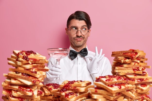 Free photo elegant pensive waiter touches bowtie, dressed in snow white uniform, suggests to degustate new cocktail, looks aside thoughtfully, poses near pile of delicious bread toasts. bartender at work