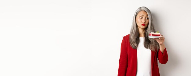 Free photo elegant senior woman in red blazer wants take bite of sweet cake looking with tempted face at desser