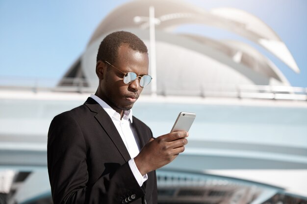 Elegant serious African American employee on business trip checking e-mail on mobile phone, standing outside modern building of airport while waiting for taxi car outdoors on sunny summer day