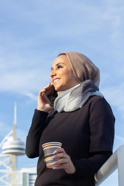 휴대 전화와 함께 우아한 미소 아랍어 여자입니다. 머리를 덮고 화장을 하고 일회용 커피 한 잔을 들고 휴대전화로 통화하는 여자. 국제, 아름다운, 소셜 미디어 개념
