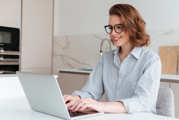メガネとストライプのシャツでエレガントな笑顔の女性が台所のテーブルに座っている間ラップトップコンピューターを使用して