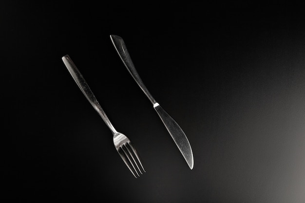 Elegant stainless steel knife and fork lying on smooth black table parallel to each other facing the viewer