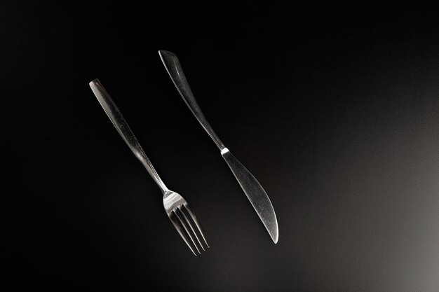 Elegant stainless steel knife and fork lying on smooth black table parallel to each other facing the viewer