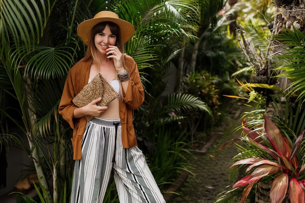 Elegant stylish girl in white top and straw hat posing on palm leaves in Bali.