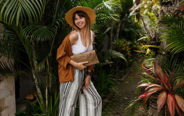 Elegant stylish girl in white top and straw hat posing on palm leaves in Bali.