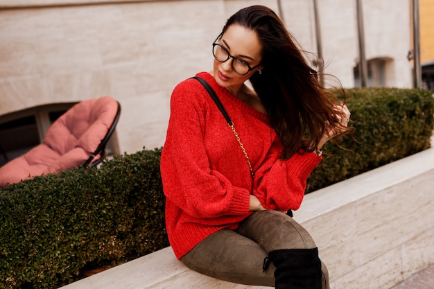 Elegant successful brunette female posing outdoor in trendy spring outfit. Fashionable boots, red knitted sweater. Old European city.