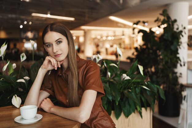 Элегантная задумчивая молодая соблазнительная женщина сидит в одиночестве в кафе, опираясь на ладонь, глядя в камеру с серьезным взглядом.