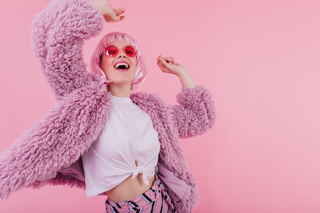 Free photo elegant white girl in pink sunglasses and peruke dancing . lovely female model posing in fur jacket and laughing