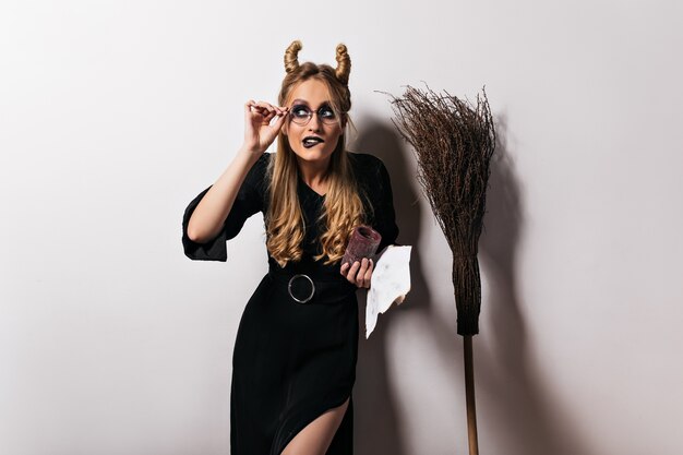 Elegant wizard in glasses funny posing on white wall. fair-haired witch standing near broom.