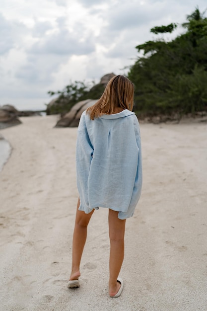 뒤에서 흐린 xAweather 보기에서 외로운 해변에서 포즈를 취하는 파란색 캐주얼 복장을 한 우아한 여성