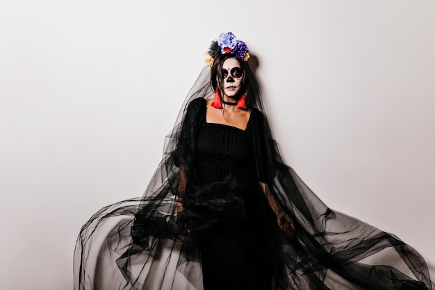 Элегантная женщина в костюме мертвой невесты с интересом смотрит в камеру Довольная модель в черном платье и вуали, стоящая на белом фоне в Хэллоуин