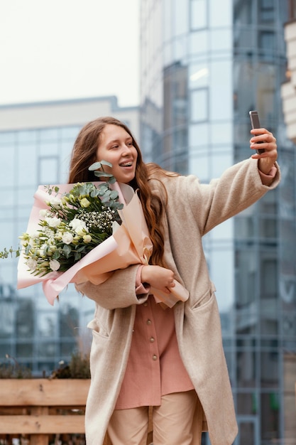 Elegant woman holding bouquet of flowers outside and taking selfie with smartphone