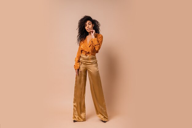 Elegant woman in orange blouse  and golden silk pants   posing  over beige wall. Hight heels. Amazing wavy hairs. full lenght.