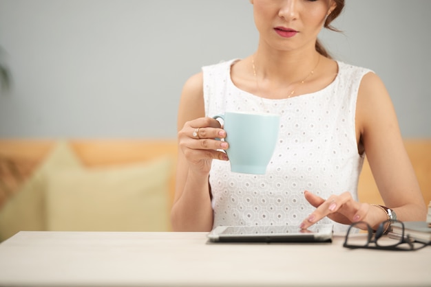 Elegant woman sitting at table indoors, using tablet and drinking from mug