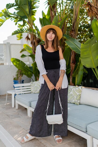 Elegant woman in straw hat posing  in cozy tropical resort near pool.