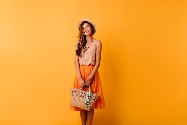 休暇の準備をしている夏服のエレガントな女性。バッグとオレンジ色にポーズをとる麦わら帽子のロマンチックな生姜の女の子。