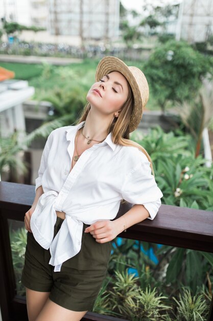 An elegant woman in a sundress stands on the balcony leaning on the railing and a straw hat on her head