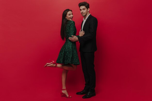 Elegant young couple in green dress and black suit touching each other smiling and posing against plain red background