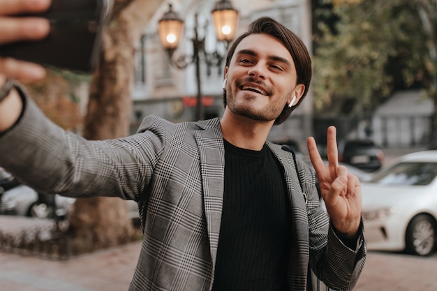 Elegant young dark-haired man in black shirt and trendy grey blazer, holding phone and making selfie against autumn city wall