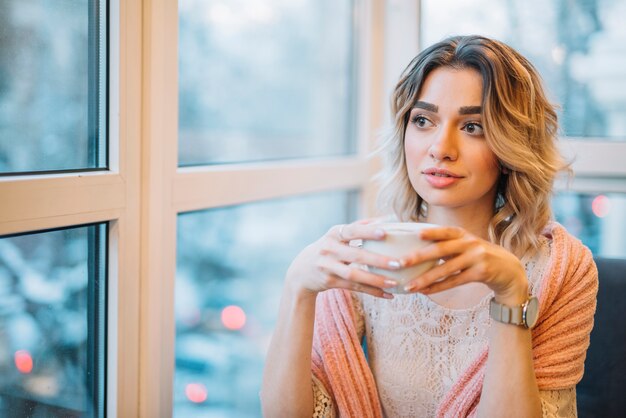 Elegant young thoughtful woman with cup of drink near window in cafe