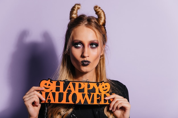 Elegante giovane strega in posa sulla parete viola. ragazza bionda in costume da vampiro che gode di halloween.