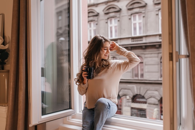 Elegant young woman in beige shirt sitting on sill and looking at city