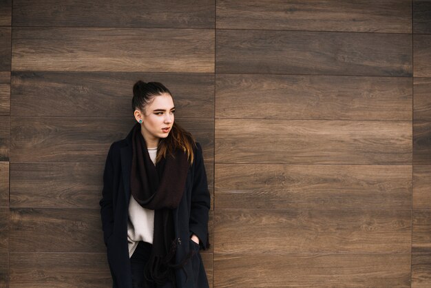 Elegant young woman in coat with scarf near wooden wall