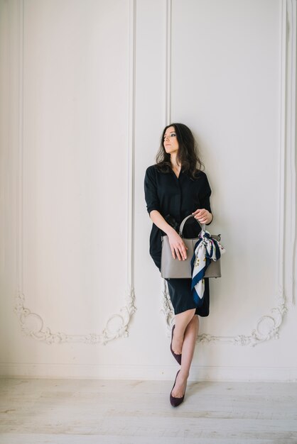 Elegant young woman in dress with handbag near wall in room