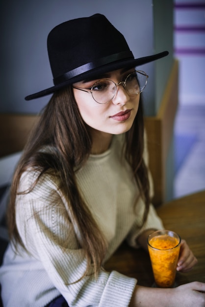 Elegant young woman in hat and eyeglasses with glass of beverage