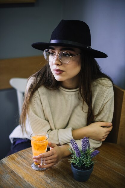 Elegant young woman in hat and eyeglasses with glass of drink and plant