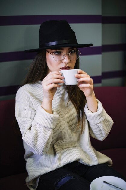 Elegant young woman in hat and eyeglasses with mug of drink