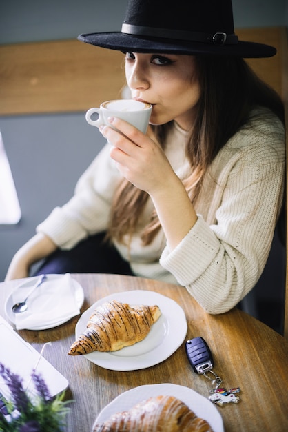 Elegant young woman in hat with mug of drink at table in cafe