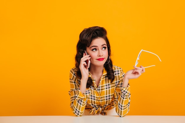 Elegant young woman talking on phone on yellow background. studio shot of wonderful pinup girl holding glasses