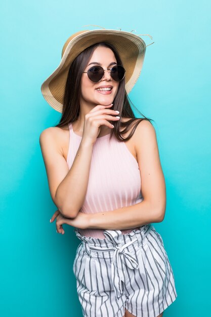 그녀의 여름 휴가에 대해 생각하고 여름 드레스, 밀짚 모자와 선글라스를 착용하는 우아한 젊은 여자. 파스텔 블루 벽 위에 절연 턱에 손 가진 여자의 측면보기.