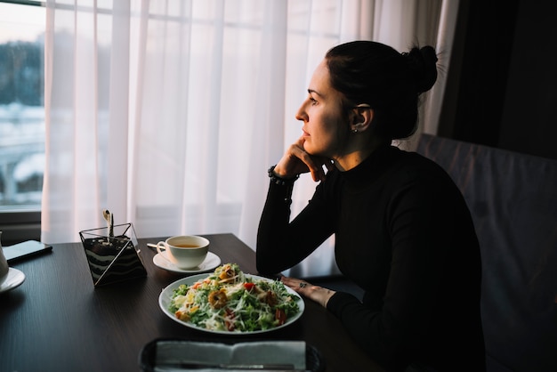 Elegant young woman with salad and cup of drink at table